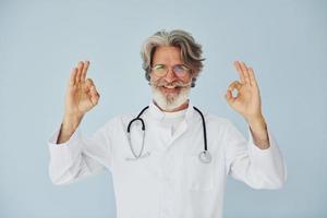 médico de jaleco branco contra a parede. homem moderno elegante sênior com cabelos grisalhos e barba dentro de casa foto