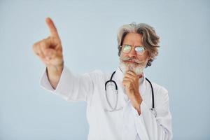 mostrando algo pelo dedo. trabalhador médico no casaco. homem moderno elegante sênior com cabelos grisalhos e barba dentro de casa foto