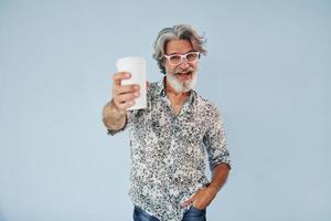 copo de bebida nas mãos. homem moderno elegante sênior com cabelos grisalhos e barba dentro de casa foto