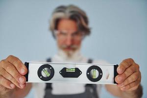 trabalhador de uniforme mantém o dispositivo de correspondência de nível. homem moderno elegante sênior com cabelos grisalhos e barba dentro de casa foto