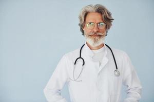 médico de jaleco branco contra a parede. homem moderno elegante sênior com cabelos grisalhos e barba dentro de casa foto