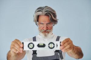 trabalhador de uniforme mantém o dispositivo de correspondência de nível. homem moderno elegante sênior com cabelos grisalhos e barba dentro de casa foto