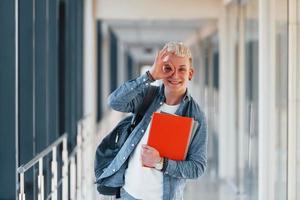 jovem estudante do sexo masculino em roupas jeans está no corredor de uma faculdade com o bloco de notas nas mãos foto