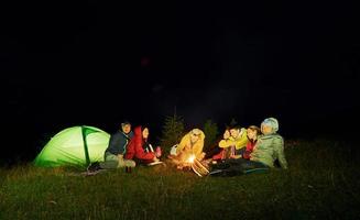 grupo de pessoas sentadas perto da fogueira e da barraca. em roupas quentes. ao ar livre durante a noite foto