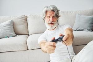 joga videogame usando o controlador. homem moderno elegante sênior com cabelos grisalhos e barba dentro de casa foto