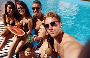 segurando a melancia. grupo de jovens felizes se divertem na piscina durante o dia foto