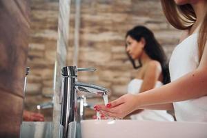 mulher jovem e bonita em pé no banheiro perto do espelho e lavando as mãos. amigos atrás foto