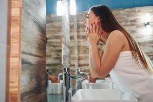 mulher jovem e bonita parada no banheiro perto do espelho e lavando o rosto foto