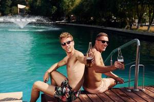 dois amigos do sexo masculino sentam-se perto da água ao ar livre com bebidas nas mãos foto