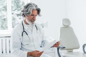 retrato do médico masculino sênior com cabelos grisalhos e barba no jaleco branco está dentro de casa na clínica foto