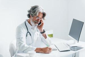 escreve pela mesa do computador. médico masculino sênior com cabelos grisalhos e barba no jaleco branco está dentro de casa na clínica foto