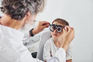 oftalmologista masculino sênior com cabelos grisalhos e barba em jaleco branco está dentro de casa na clínica testando a visão de uma garotinha foto