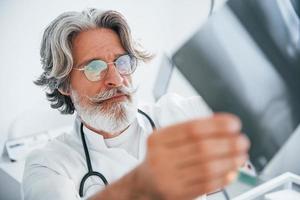 olha para o raio-x. médico masculino sênior com cabelos grisalhos e barba no jaleco branco está dentro de casa na clínica foto