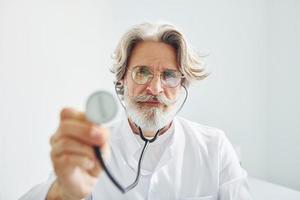 segura o estetoscópio. retrato do médico masculino sênior com cabelos grisalhos e barba no jaleco branco está dentro de casa na clínica foto