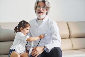 menina brincando com estetoscópio. médico masculino sênior com cabelos grisalhos e barba no jaleco branco está dentro de casa na clínica foto