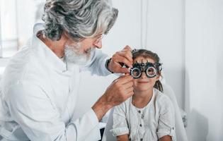 oftalmologista masculino sênior com cabelos grisalhos e barba em jaleco branco está dentro de casa na clínica testando a visão de uma garotinha foto