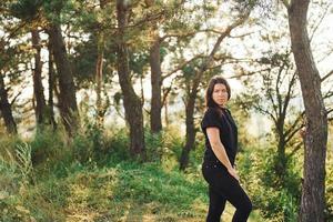 morena de camisa preta e jeans em pé e posando na floresta durante o dia com o sol lindo foto