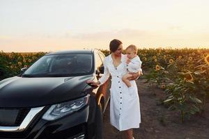 perto de carro preto moderno. jovem mãe com seu filho pequeno está ao ar livre no campo agrícola. lindo sol foto