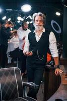 de pé e fumando na barbearia. elegante homem sênior moderno com cabelos grisalhos e barba está dentro de casa foto