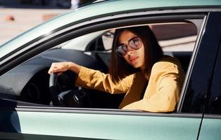 proprietário de automóvel feliz senta-se dentro do veículo. jovem elegante em casaco cor de vinho durante o dia com seu carro foto