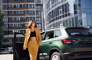 caminha para a frente do automóvel. jovem elegante em casaco cor de vinho durante o dia com seu carro foto