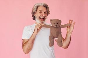 segura o ursinho de pelúcia. elegante homem sênior moderno com cabelos grisalhos e barba está dentro de casa foto