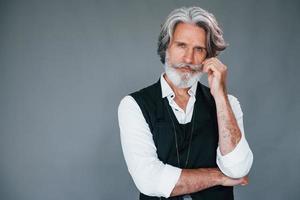 contra um fundo cinza. elegante homem sênior moderno com cabelos grisalhos e barba está dentro de casa foto