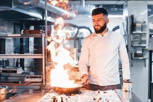 frigideira está pegando fogo. chef de uniforme branco cozinhando comida na cozinha. dia ocupado no trabalho foto