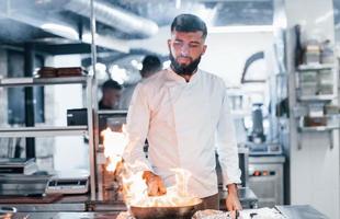 frigideira está pegando fogo. chef de uniforme branco cozinhando comida na cozinha. dia ocupado no trabalho foto