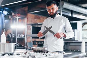 chef de uniforme branco em pé na cozinha. segurando facas nas mãos foto