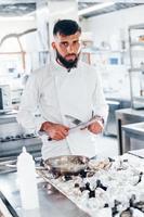 chef de uniforme branco em pé na cozinha. segurando facas nas mãos foto