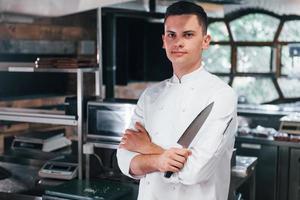 chef de uniforme branco em pé na cozinha. segurando a faca nas mãos foto