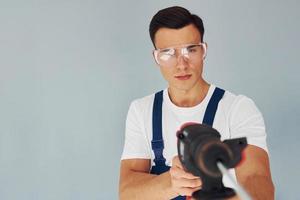 em óculos de proteção e com broca nas mãos. trabalhador do sexo masculino em pé uniforme azul dentro do estúdio contra fundo branco foto