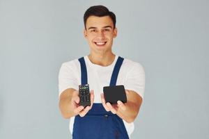 controle remoto de tv e smartphone. trabalhador do sexo masculino em pé uniforme azul dentro do estúdio contra fundo branco foto