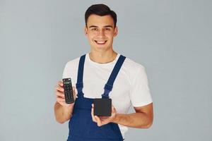 controle remoto de tv e smartphone. trabalhador do sexo masculino em pé uniforme azul dentro do estúdio contra fundo branco foto