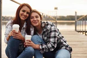 lindo casal de lésbicas senta-se perto do lago com copos de bebida nas mãos e se abraçando foto