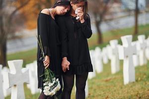 segura flores. duas jovens em roupas pretas visitando o cemitério com muitas cruzes brancas. concepção de funeral e morte foto