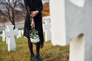 com flores nas mãos. jovem mulher em roupas pretas, visitando o cemitério com muitas cruzes brancas. concepção de funeral e morte foto