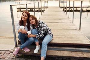 lindo casal de lésbicas senta-se perto do lago com copos de bebida nas mãos e se abraçando foto