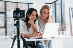 câmera no tripé está gravando. duas jovens freelancers femininas trabalhando dentro de casa no escritório com laptop foto