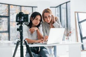 câmera no tripé está gravando. duas jovens freelancers femininas trabalhando dentro de casa no escritório com laptop foto