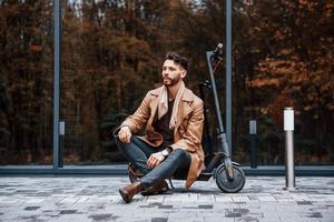 contra a construção e com scooter elétrico. jovem modelo masculino em roupas da moda está ao ar livre na cidade durante o dia foto