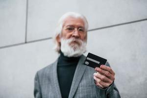 empresário sênior em roupas formais, com cabelos grisalhos e barba está ao ar livre detém cartão de crédito foto