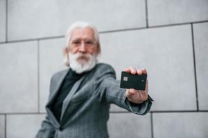 empresário sênior em roupas formais, com cabelos grisalhos e barba está ao ar livre detém cartão de crédito foto