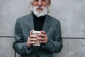 empresário sênior em roupas formais, com cabelos grisalhos e barba está ao ar livre e segura um copo de bebida foto