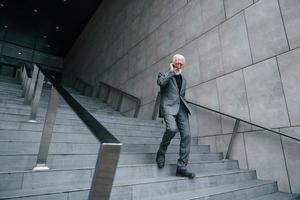 empresário sênior em roupas formais, com cabelos grisalhos e barba é falar ao ar livre por telefone foto