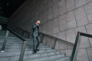 empresário sênior em roupas formais, com cabelos grisalhos e barba é falar ao ar livre por telefone foto