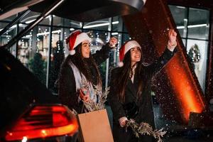 parado perto do porta-malas do carro aberto. irmãs gêmeas felizes passam as férias de natal juntas ao ar livre. concepção de ano novo foto