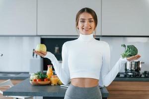 posa para uma câmera com legumes. jovem europeia está dentro de casa na cozinha dentro de casa com comida saudável foto