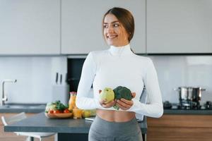 posa para uma câmera com legumes. jovem europeia está dentro de casa na cozinha dentro de casa com comida saudável foto